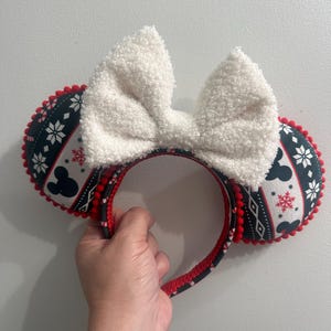 May include: A festive headband featuring Mickey Mouse ears. The ears are black with a white snowflake and Mickey Mouse pattern, trimmed with red pom-poms. A large, fluffy white bow sits atop the headband. The headband is held by a hand.