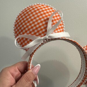 May include: Orange and white gingham pattern mouse ear headband with a white satin bow. The ears are outlined with small white pearls. The headband is trimmed with white lace and gingham fabric.