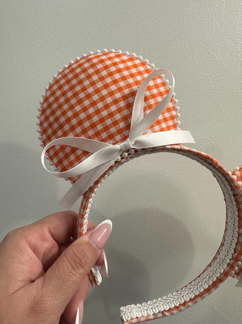 May include: Orange and white gingham pattern mouse ear headband with a white satin bow. The ears are outlined with small white pearls. The headband is trimmed with white lace and gingham fabric.