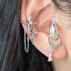 Puede incluir: Juego de puño de oreja y pendientes de aro plateados. El puño de oreja presenta un diseño de estrella con una cadena colgante. Los pendientes de aro tienen un pequeño dije colgando. La joyería se usa en la oreja.