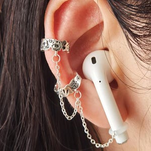 Puede incluir: Joyería de plata con puño y cadena para la oreja. El puño para la oreja presenta un diseño decorativo y está conectado a un segundo puño con una cadena. Un auricular blanco está en la oreja.