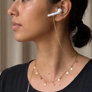 May include: A gold-toned metal chain accessory designed to secure wireless earbuds, featuring a delicate link chain adorned with small, evenly spaced star-shaped charms. The chain connects to a white earbud and drapes around the neck like a necklace.