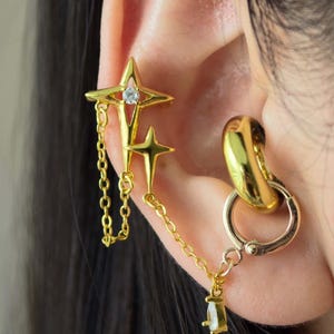 Puede incluir: Joyería dorada para la oreja con un brazalete en forma de estrella con una cadena colgante y una estrella pequeña, conectada a un pendiente de aro. La joyería se usa en la oreja, con un enfoque en el diseño intrincado y los detalles.