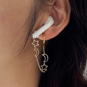 Puede incluir: Pendientes dorados con un diseño celestial de estrellas y lunas crecientes. Los pendientes están conectados por una delicada cadena y cuelgan de un aro dorado. Un auricular blanco está en la oreja.