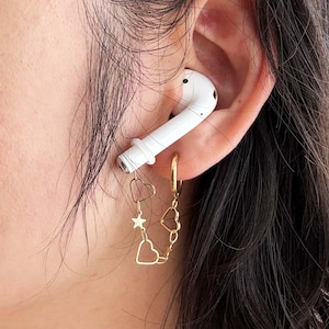 Pode incluir: Um brinco dourado com uma corrente de corações e uma estrela é preso a um fone de ouvido branco. O brinco é conectado a um brinco de argola dourado. O fone de ouvido está na orelha de uma pessoa com cabelo escuro.