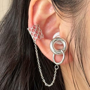 Puede incluir: Puño de oreja plateado y pendientes de doble aro conectados por una delicada cadena. El puño presenta un diseño de panal, mientras que los pendientes tienen dos círculos entrelazados. La joya se lleva en la oreja.