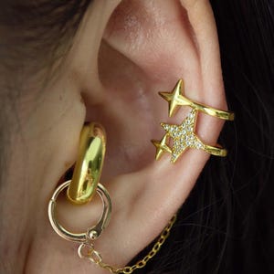 Puede incluir: Juego de aretes dorados con un arete de aro con cadena y un brazalete de oreja en forma de estrella. El brazalete de oreja tiene un diseño de doble banda con una estrella y un grupo de pequeños cristales. Las joyas se usan en la oreja.