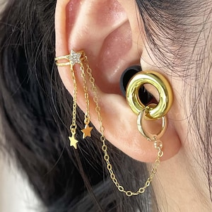 May include: Gold-toned ear cuff and earring set. The ear cuff features a star design with dangling chains and star charms. The earring is a large gold hoop with a smaller hoop attached, connected by a chain.