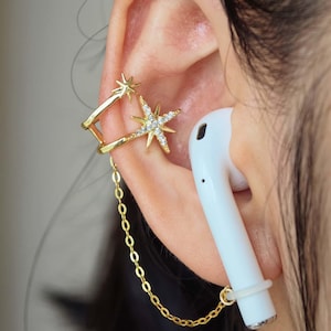 Puede incluir: Puño de oreja dorado con diseños de estrellas y estrellas, conectado por una delicada cadena a un auricular blanco. El puño de oreja presenta pequeños acentos brillantes. El auricular está asegurado con un anillo de silicona blanco.
