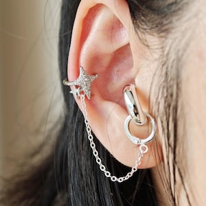 Puede incluir: Un conjunto de pendientes y puño de oreja plateados. El puño de oreja presenta un diseño de estrella con pequeñas piedras transparentes. Una cadena conecta el puño de oreja con un pendiente de aro plateado.