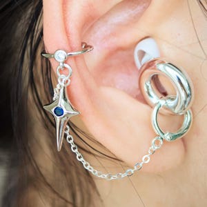 Puede incluir: Puño de oreja plateado con un dije de estrella con una piedra azul en el centro, conectado por una cadena a un pendiente de doble aro. El puño de la oreja tiene una pequeña piedra transparente.