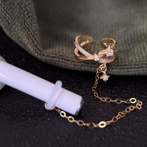 Puede incluir: Un pendiente de oreja dorado con una delicada cadena y dijes de estrella está unido a un auricular inalámbrico blanco. El pendiente de oreja presenta un diseño entrecruzado y está adornado con pequeñas piedras preciosas transparentes. El fondo es una superficie oscura y tela verde.