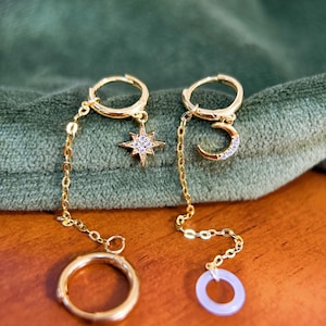 May include: Gold earrings with celestial motifs. One earring has a star pendant with clear stones, the other a crescent moon with similar stones. Both dangle from a hoop and chain, with a ring charm at the bottom.