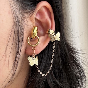 Puede incluir: Juego de puño y pendientes de color dorado. El puño presenta un diseño de mariposa, y el pendiente tiene un aro con un dije de mariposa conectado por una delicada cadena. La joya se lleva en la oreja.