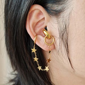 Puede incluir: Juego de pendientes y puño de oreja de color dorado. El puño de oreja presenta un diseño de aro grande, mientras que los pendientes incluyen una cadena de pequeños abalorios de estrella. La joya se lleva en la oreja, con la cadena de estrellas colgando.