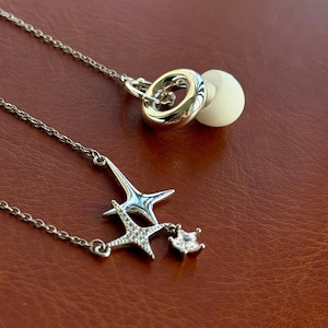 May include: A silver-toned necklace featuring a delicate chain with a pendant composed of two layered star shapes, one with a textured surface and one with a clear stone, plus a small dangling star charm and a circular earbud attachment.