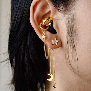 Könnte beinhalten: Ein goldfarbenes Ohrklemmen- und Ohrring-Set, bestehend aus einer glatten Ringklemme, die über feine Ketten mit einem kleinen Stern, einem Halbmond und einem weiteren Stern verbunden ist. Ein separater Stern-Ohrstecker mit klaren Steinen ist dabei.