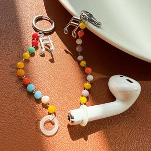 May include: A white wireless earbud with a colorful beaded chain and musical note charms. The chain features a variety of colorful beads, including red, orange, yellow, blue, and white. The earbud is resting on a brown surface.