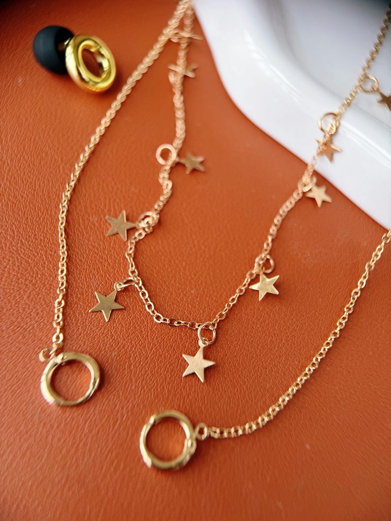May include: A gold-toned chain necklace featuring multiple small star-shaped charms suspended along the links. The necklace includes circular ring connectors at the ends and is accompanied by a separate gold-toned hoop earring with a black backing.
