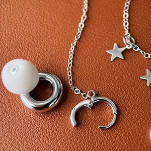 May include: A silver-toned jewelry set consisting of a hoop earring with a white spherical bead attachment, and a delicate chain featuring multiple small star-shaped charms connected to a hinged hoop earring component.