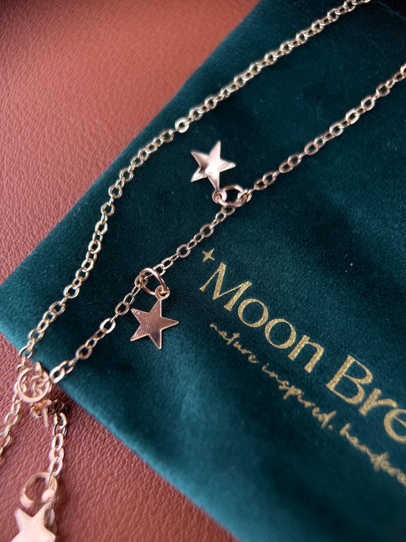 May include: A gold-toned metal chain necklace featuring multiple small, flat, five-pointed star charms dangling from the links. The necklace is positioned over a dark green velvet pouch with gold text that reads Moon Bre and nature inspired.