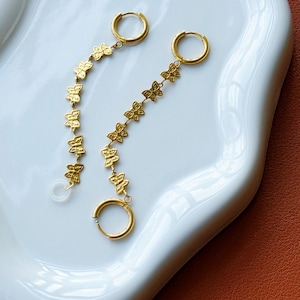Könnte beinhalten: Ein Paar goldfarbene Creolen mit hängendem Kettendesign. Jeder Ohrring besteht aus einem runden Ring, der mit einer vertikalen Kette aus kleinen, sich wiederholenden schmetterlingsförmigen Anhängern verbunden ist, mit einem transparenten Kunststoffteil am Ende.