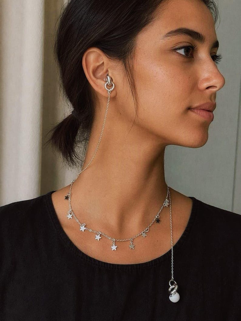 May include: A silver-toned ear cuff connected by a delicate chain to a necklace featuring multiple small star charms. The chain extends from the ear cuff down to the collarbone area, where it joins a necklace adorned with star-shaped pendants.