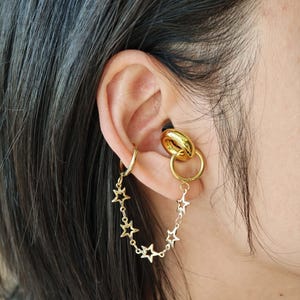 May include: Gold-colored ear jewelry set. The set includes a small hoop earring, a double hoop earring, and a chain of star-shaped charms. The jewelry is worn on the ear, with the chain of stars dangling down.