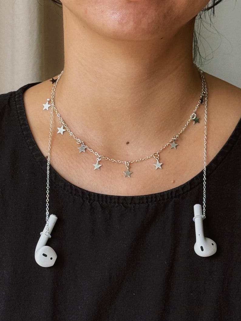 May include: A silver-toned metal chain necklace featuring multiple small star-shaped charms hanging along the length. The chain includes two integrated white plastic earbud holders at the ends, designed to secure wireless earbuds while worn.