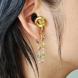 May include: A gold-toned ear cuff and hoop earring set connected by a delicate chain. The chain features small, round, multi-colored gemstones in shades of blue, pink, green, and orange. The jewelry is worn in the ear, with a black earplug visible.