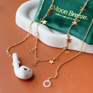 May include: A gold-toned chain accessory for wireless earbuds featuring small star-shaped charms attached at intervals. The chain includes a white silicone ring at one end and connects to a white wireless earbud at the other end, with a green pouch.