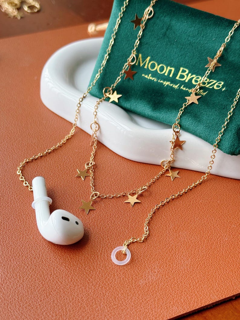 May include: A gold-toned chain accessory for wireless earbuds featuring small star-shaped charms attached at intervals. The chain includes a white silicone ring at one end and connects to a white wireless earbud at the other end, with a green pouch.