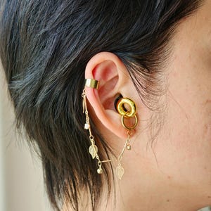 Puede incluir: Un pendiente tipo ear cuff dorado conectado por una cadena fina a un aro dorado. La cadena presenta pequeños dijes en forma de hoja y diminutos cristales transparentes. El aro está insertado en un expansor negro para la oreja.