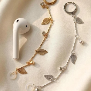 May include: Gold and silver earbud chains with leaf and crystal accents. One gold chain is paired with a white earbud. The chains feature a delicate design with small leaves and sparkling crystals, perfect for accessorizing.