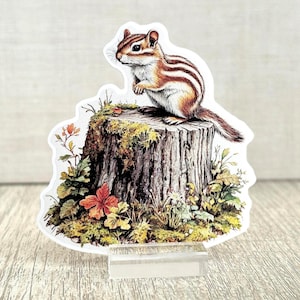 May include: A sticker featuring a chipmunk with brown and white stripes, perched atop a tree stump covered in moss and surrounded by colorful foliage. The sticker is mounted on a clear acrylic stand.