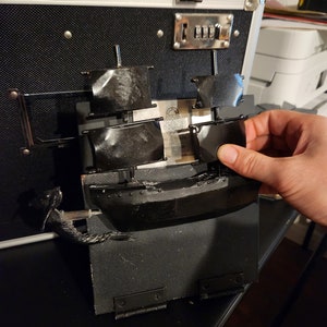 Op de afbeelding: Een zwart houten scheepsmodel met zeilen, gemonteerd op een zwarte houten basis met scharnieren. Het schip wordt vastgehouden door een hand.