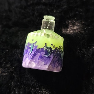 May include: A small, decorative bottle with a clear stopper. The bottle is filled with a mix of green and purple glitter and has the number "5" on the side. The bottle is sitting on a black surface.
