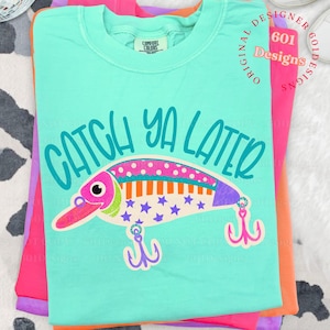May include: A turquoise Comfort Colors t-shirt with the phrase "CATCH YA LATER" and a colorful cartoon fishing lure graphic. The lure features pink, orange, and purple accents with star details. The shirt is folded with other brightly colored shirts.