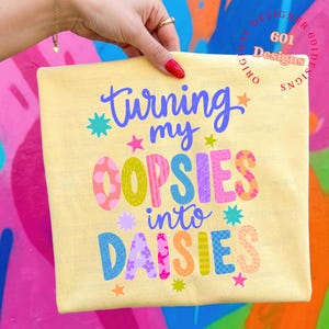 May include: A light yellow t-shirt with the phrase "Turning my OOPSIES into DAISIES" in colorful, playful lettering. The design includes stars and floral accents, creating a cheerful and whimsical aesthetic.