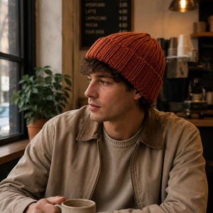 Puede incluir: Un gorro de punto acanalado de color óxido con un borde doblado. El gorro presenta un patrón vertical texturizado en toda la tela, proporcionando un ajuste ceñido alrededor de la cabeza. El material parece ser un hilo grueso y cálido.