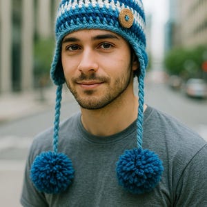 Könnte beinhalten: Blau-weiße gehäkelte Mütze mit Ohrenklappen und zwei blauen Bommeln. Die Mütze hat ein Holzknopfdetail. Ein Winteraccessoire.