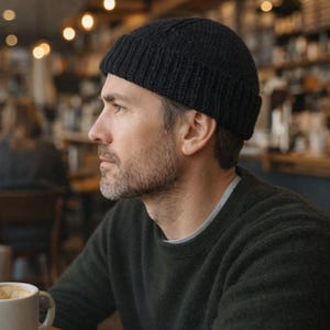 Könnte beinhalten: Eine schwarze Strickmütze. Die Mütze wird von einer Person mit Bart und grau werdendem Haar getragen. Die Person trägt einen dunkelgrünen Pullover und sitzt in einem Café.