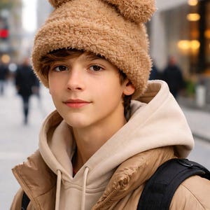 May include: A person wearing a tan teddy bear hat with two fluffy ears. The person is also wearing a tan puffer jacket and a beige hoodie. A black backpack is visible on the person's back.