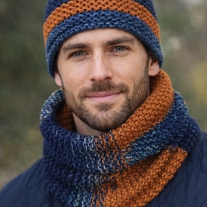 Puede incluir: Un conjunto de gorro y cuello de punto en tonos azul marino, naranja y verde azulado. El gorro presenta una franja naranja horizontal. El cuello tiene un patrón de color similar. El conjunto está hecho de hilo texturizado.
