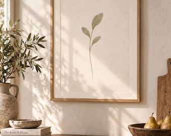 May include: A framed wall art print featuring a single, delicate green botanical leaf illustration centered on a textured off-white background. The artwork is enclosed in a simple, light-toned wooden frame, displaying a clean and neutral aesthetic.