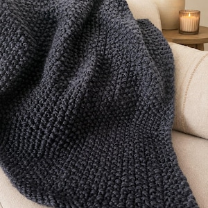 May include: A dark gray, chunky knit blanket draped over a beige sofa. The blanket's texture is prominent, with visible stitches creating a dense, warm appearance. The scene suggests comfort and relaxation, with soft lighting.