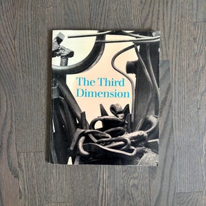 May include: A book titled "The Third Dimension" with a black and white abstract sculpture on the cover. The book is lying on a dark wood surface. The title is in a teal color.