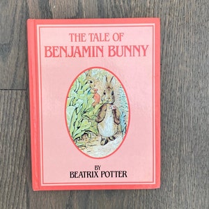 May include: A pink hardcover book with the title "The Tale of Benjamin Bunny" by Beatrix Potter. The cover features an illustration of a rabbit holding a flower.