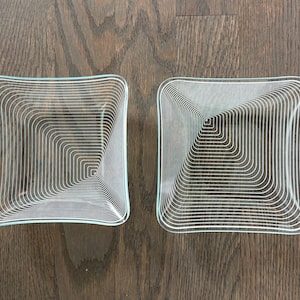 May include: Two clear glass square dishes with a white concentric square design. The dishes have slightly curved edges and are displayed on a dark wood surface. The design creates a visual illusion of depth.