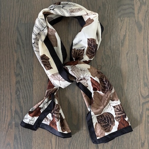 May include: A long rectangular scarf featuring a cream-colored background with a pattern of brown leaves and black fern illustrations. The scarf has a solid black border along the edges and the word OSCAR is printed in the bottom left corner.